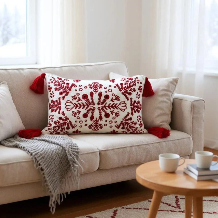 Handcrafted white cotton throw pillow with intricate red embroidery and tassels, styled on a beige sofa in a cozy winter living room with natural light.