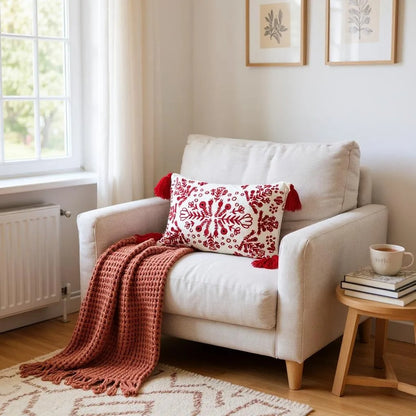Red and white embroidered boho pillow with tassels placed on a cream armchair, styled with a rust knit throw blanket in a warm, inviting corner with natural decor.