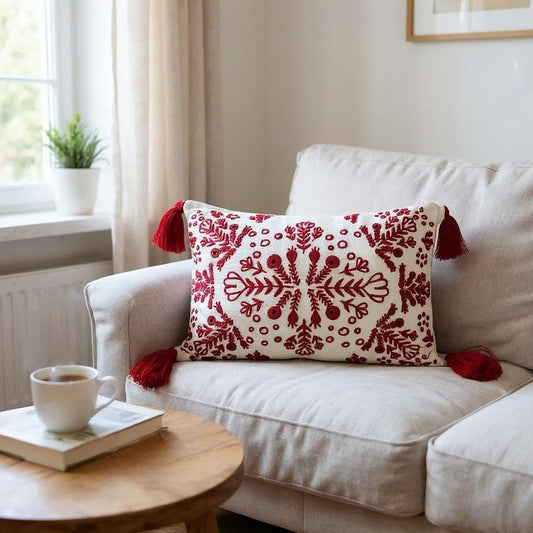 Bohemian-style rectangular pillow with red embroidery and corner tassels, displayed on a neutral armchair with a cozy knit blanket and books, creating a relaxed, stylish space.